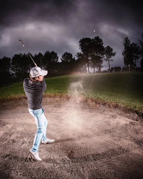 Nils Bouchilloux frappe une balle depuis un bunker, le sable projeté en gerbe, sous un ciel d'orage